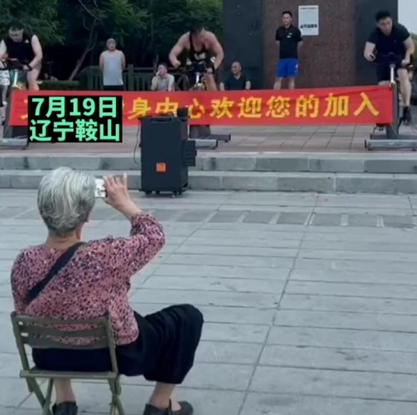 肌肉小伙在广场健身，大妈搬板凳坐对面观看，看不清楚拿手机录像